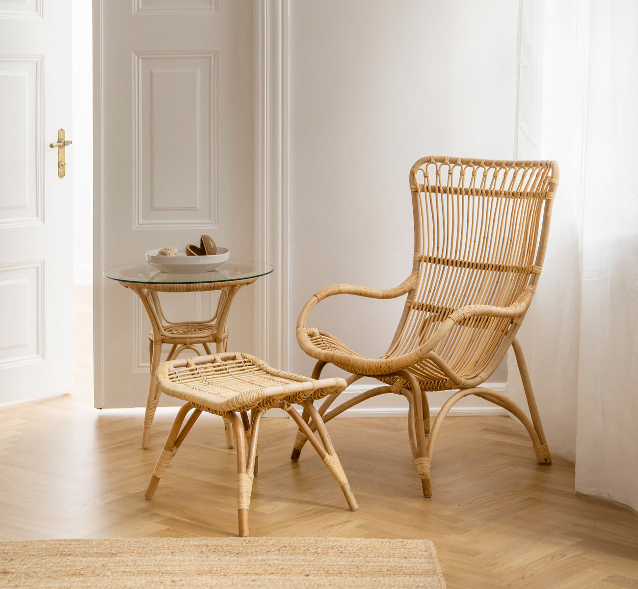 This is a lifestyle image of Monet Footstool in natural rattan. Paired with a high-back rattan chair and glass side table in a bright entry.