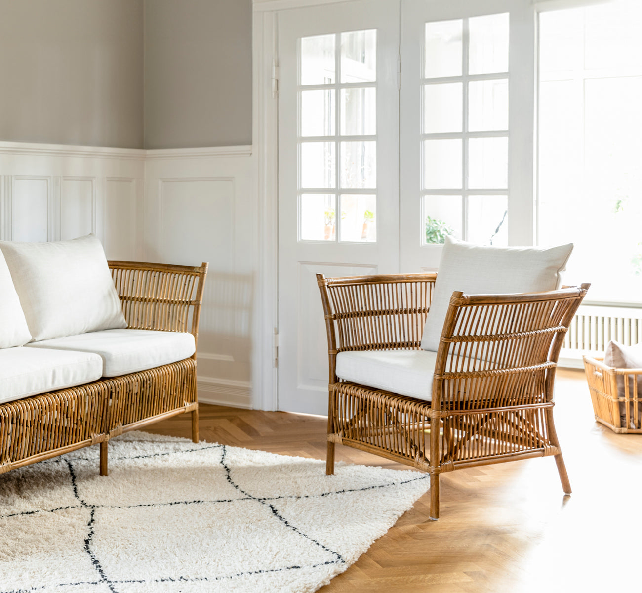 This is a lifestyle image of Donatello Lounge Chair in antique rattan, in a bright living room with herringbone parquet and lush greenery.