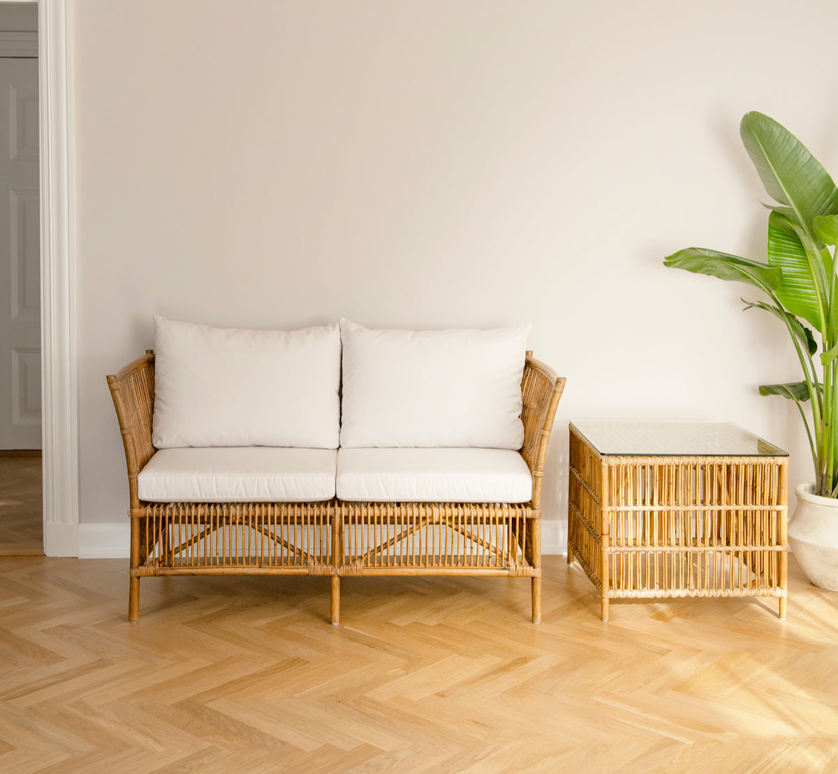 This is a lifestyle image of Donatello 2 Seater Sofa in antique rattan, in a minimal bright interior with herringbone parquet and a tropical plant.
