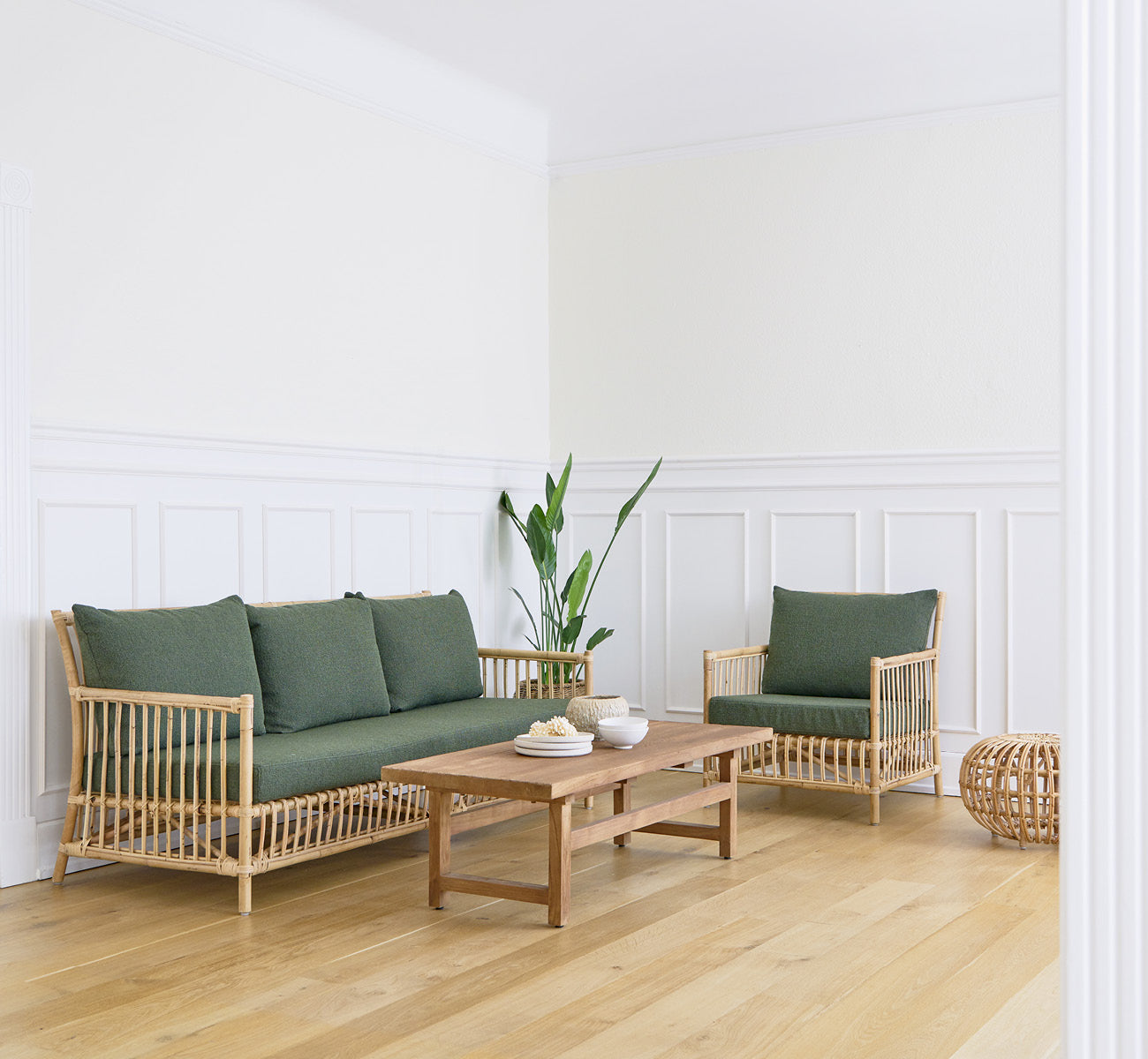 This is a lifestyle image of Caroline Sofa in natural rattan, in a bright interior with white panelled walls and herringbone parquet.