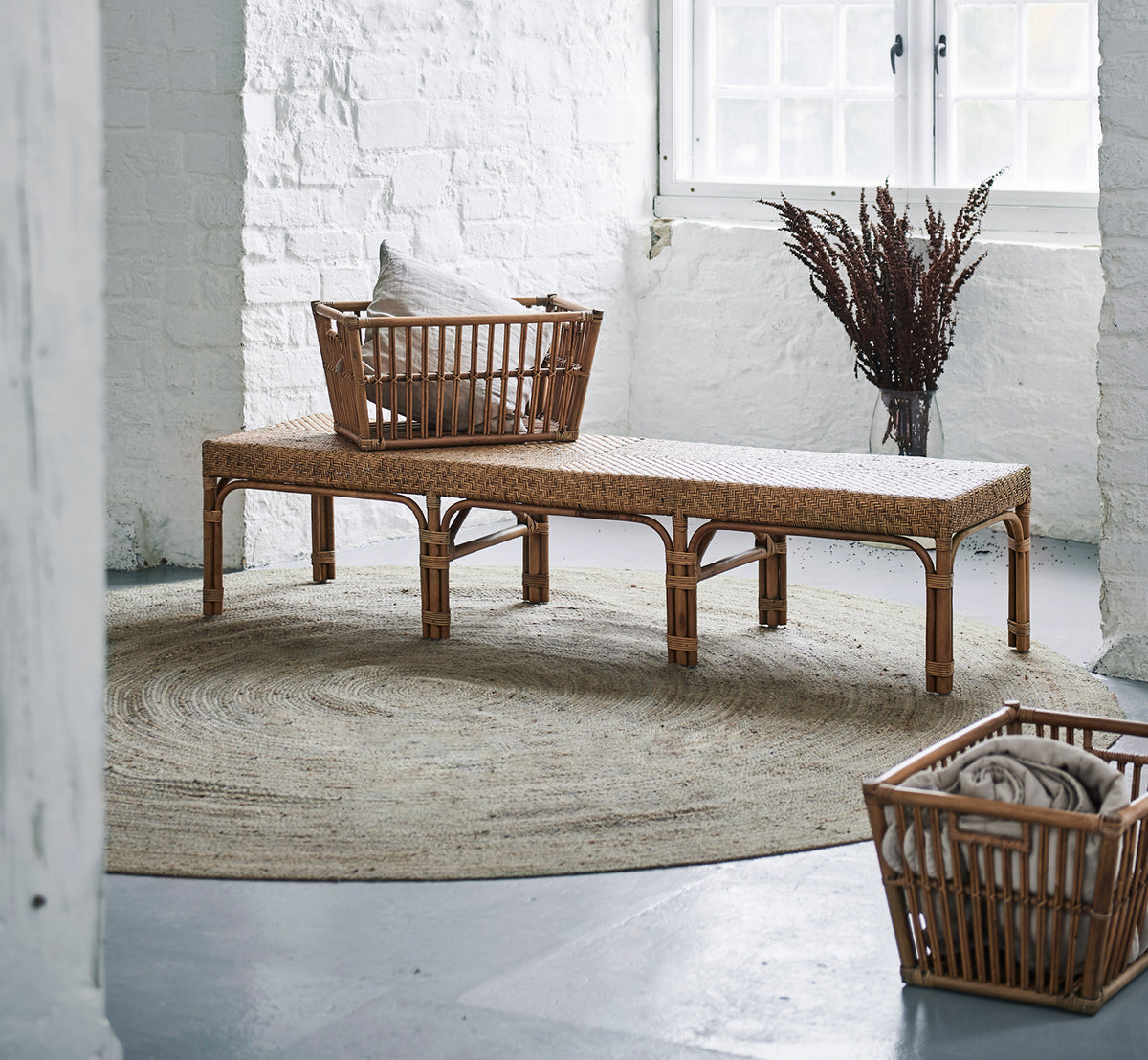 This is a lifestyle image of Luis Bench in antique rattan. Placed in a white brick loft with a basket, jute rug, and dried grasses as décor.