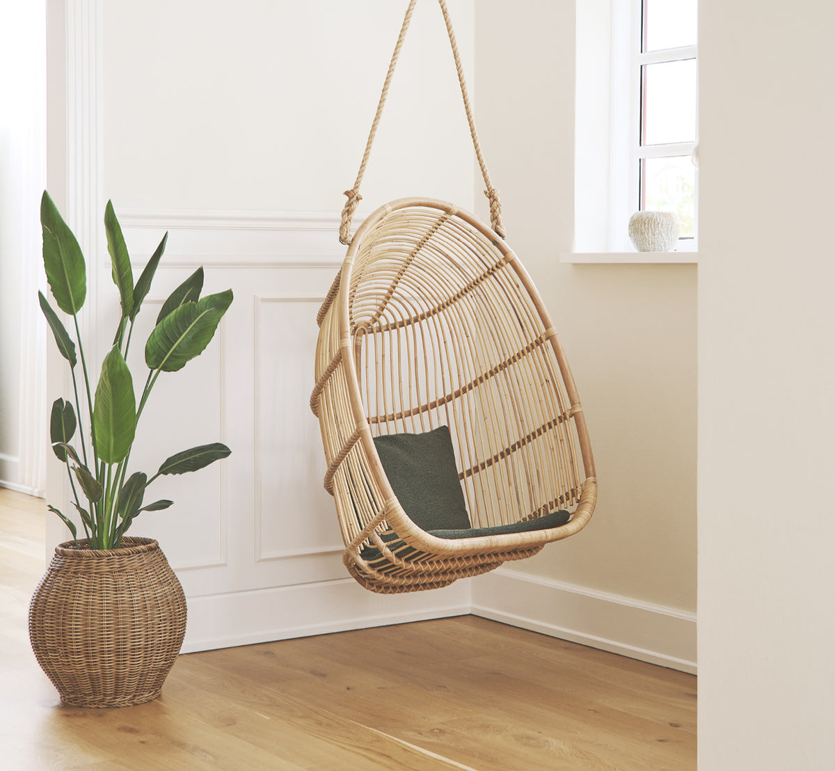 This is a lifestyle image of Renoir Hanging Chair in natural rattan. Hanging in a bright panelled entry beside a potted leaf plant.
