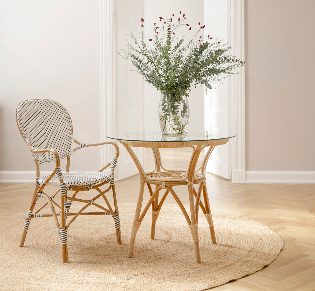 This is a lifestyle image of Tony Café Table in natural rattan. Paired with two chairs on a round jute rug in a bright doorway.