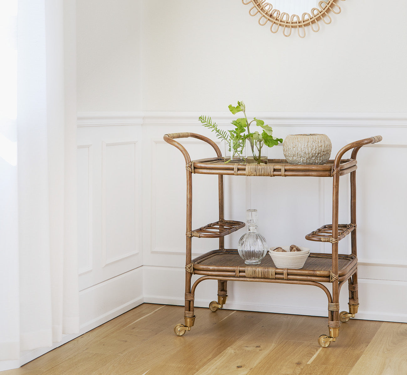 This is a lifestyle image of Carlo Trolley in antique Rattan, in a bright white interior with wainscoting and a rattan wall mirror.