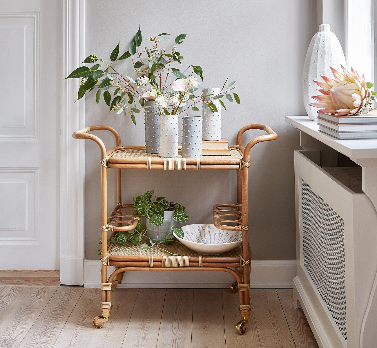 This is a lifestyle image of Carlo Trolley in antique Rattan, in a bright white interior with wainscoting and a rattan wall mirror.