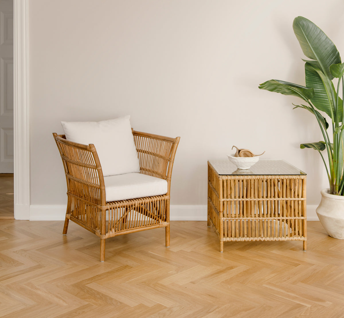 This is a lifestyle image of Donatello Side Table in antique rattan, in a bright interior with white panelled walls and herringbone parquet.