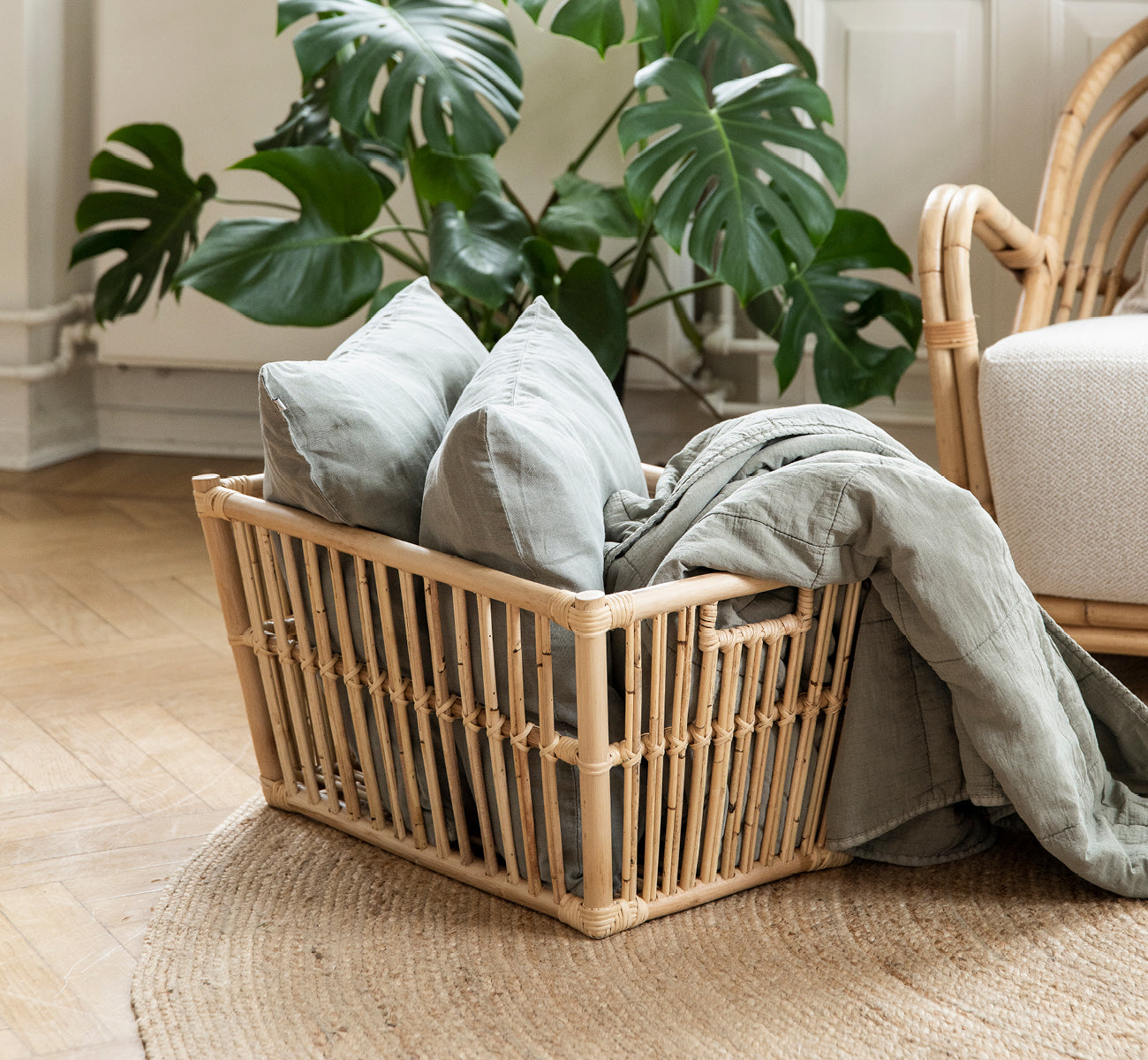This is a lifestyle image of Marche Basket in natural rattan. Filled with linen cushions beside a monstera plant on oak floor.