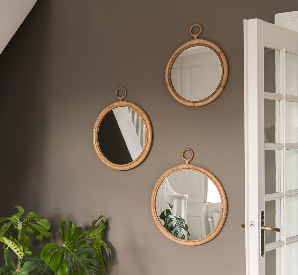 This is a lifestyle image of Ella Mirror O50cm in natural skin on rattan, on a taupe wall in a hallway with a monstera plant and white door.