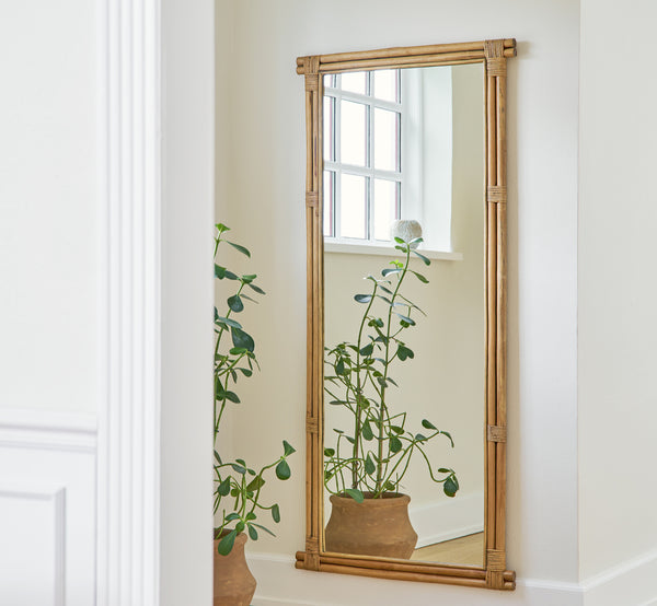 This is a lifestyle image of Rasmus Mirror 172 cm in antique rattan. Mounted on a white wall above a potted plant in a bright corridor.