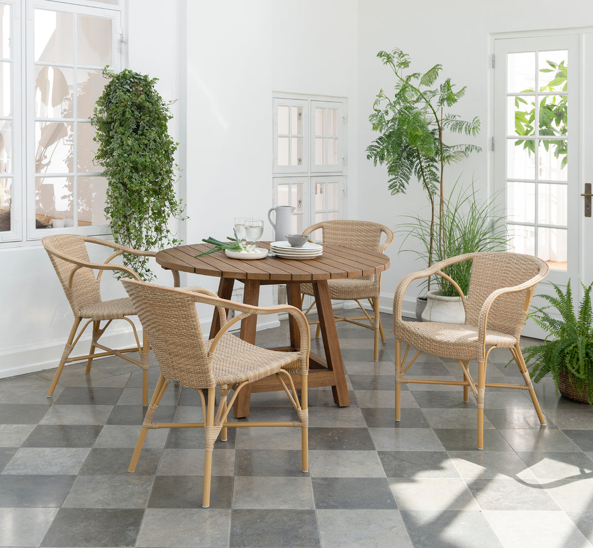 This is a lifestyle image of Madeleine Exterior Dining Chair in almond aluminium frame natural ArtFibre. Bright conservatory with plants and tiles.