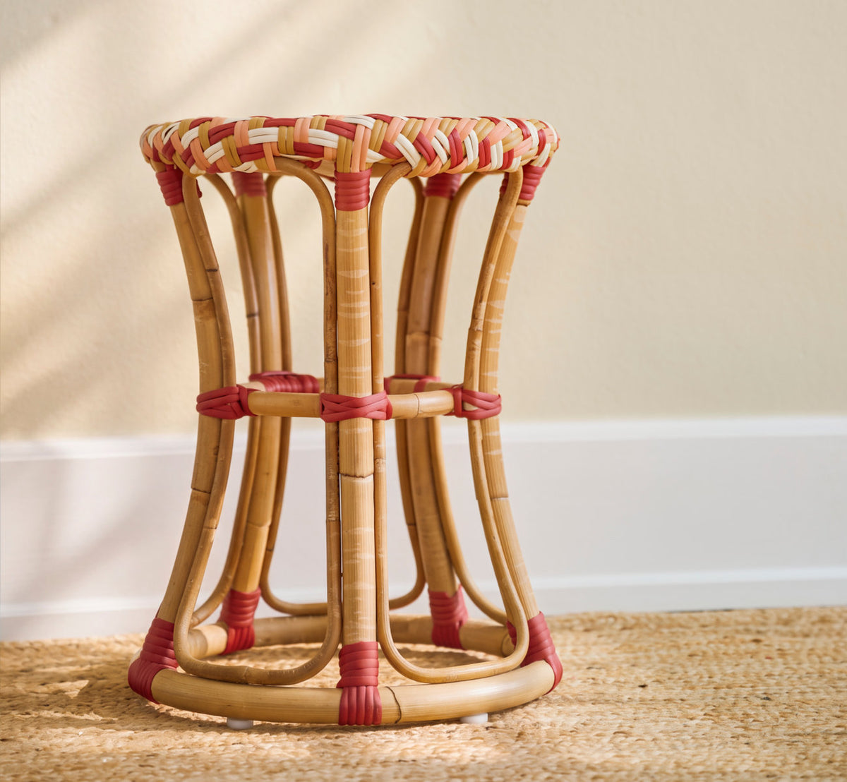 This is a lifestyle image of Chloe Stool in zigzag weave ivory honey coral burgundy red skin on rattan frame. On a jute rug against a warm cream wall.