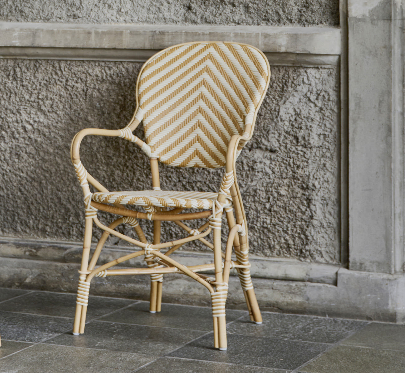 This is a lifestyle image of Isabell Dining Armchair in chevron Honey Ivory ArtFibre Skin on Rattan frame, in a bright dining area with large windows.