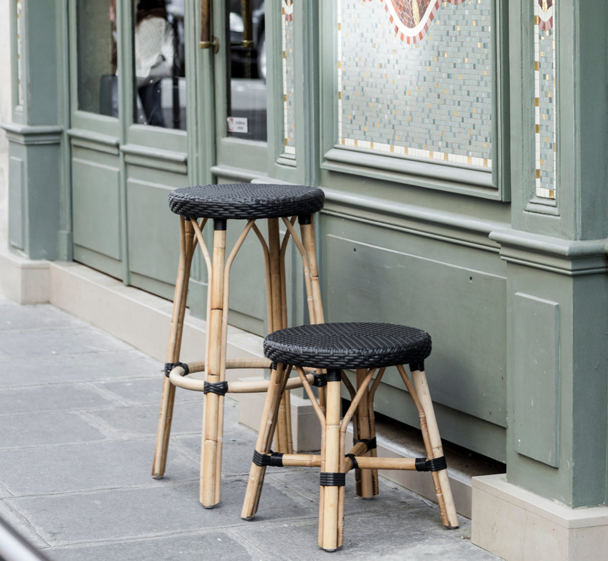 This is a lifestyle image of Simone Counter Stool in black ArtFibre Skin on Rattan frame. Placed outside a green Parisian café storefront.