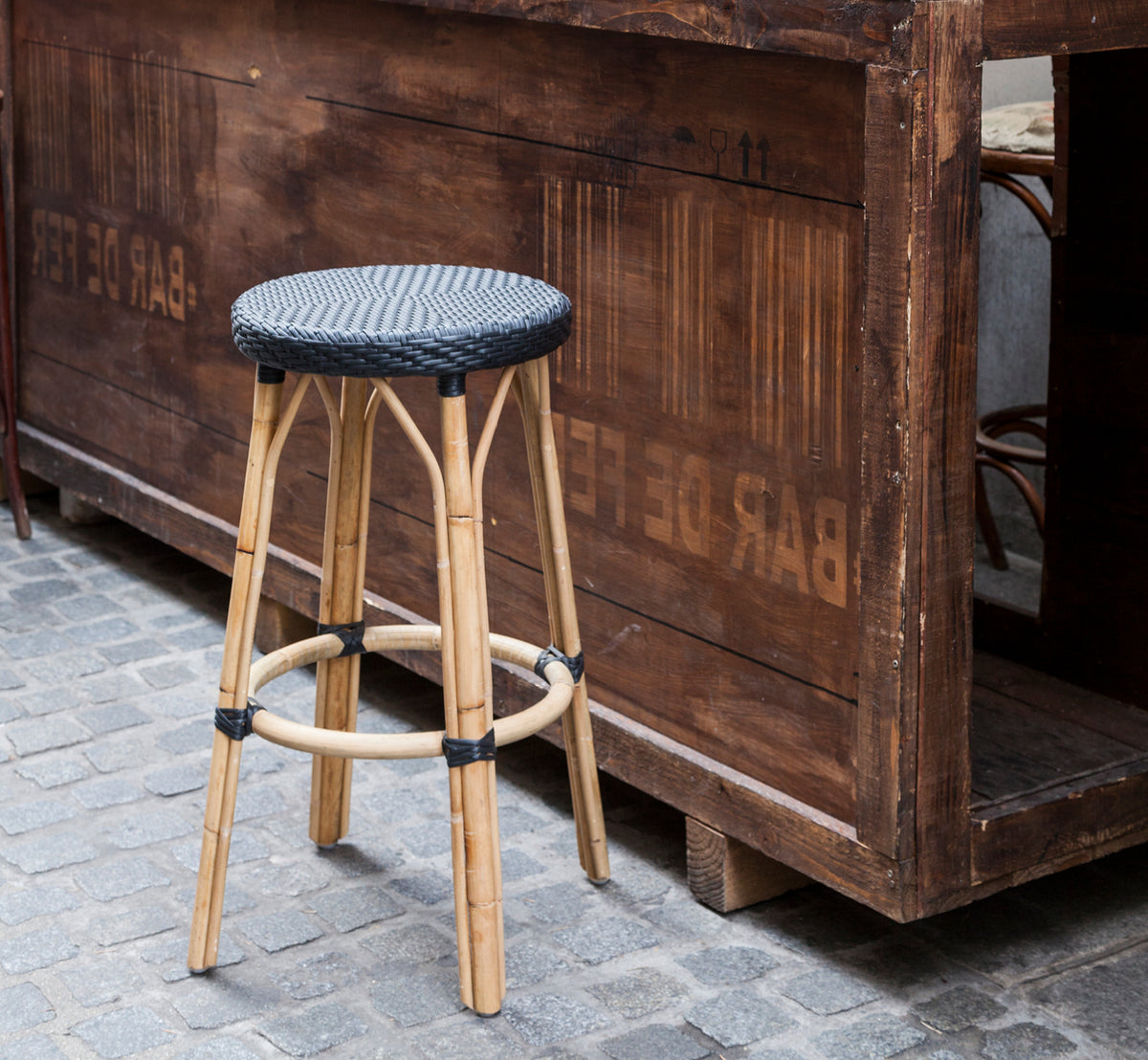 This is a lifestyle image of Simone Bar Stool in black ArtFibre Skin on Rattan frame. Placed by a rustic wooden bar on cobblestones.