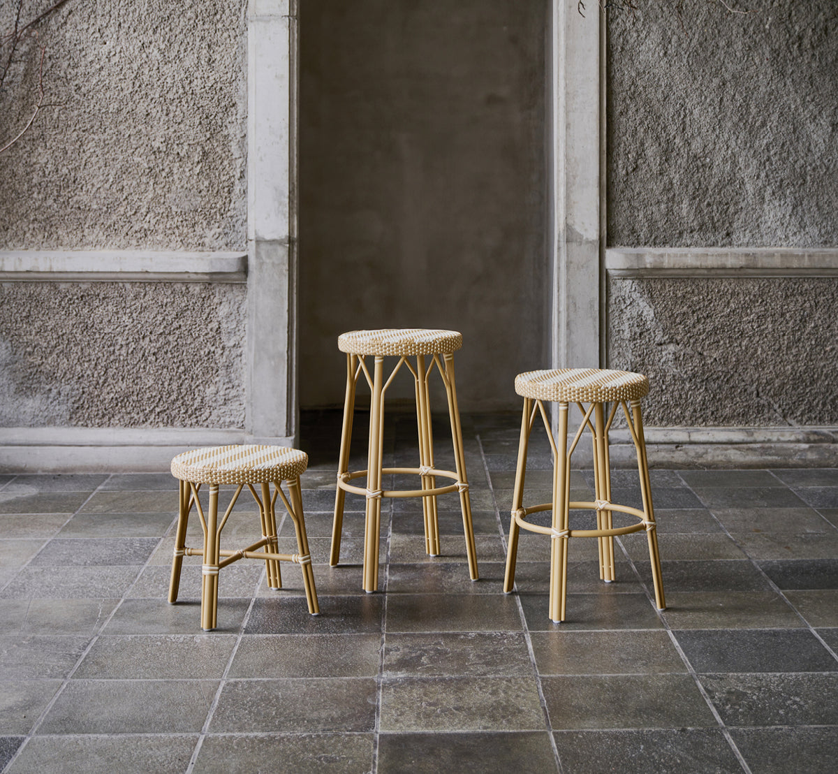 This is a lifestyle image of Simone Bar Stool in striped honey ivory ArtFibre Skin on Rattan frame. Shown in a stone courtyard.