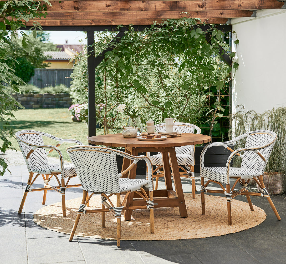 This is a lifestyle image of Madeleine Dining Chair in white w cappuccino dot ArtFibre skin on rattan frame. Garden pergola with jute rug.