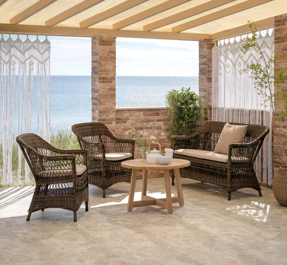 This is a lifestyle image of Charlot Exterior Chair in brown artfibre aluminum. With teak coffee table and macramé curtains by the sea.