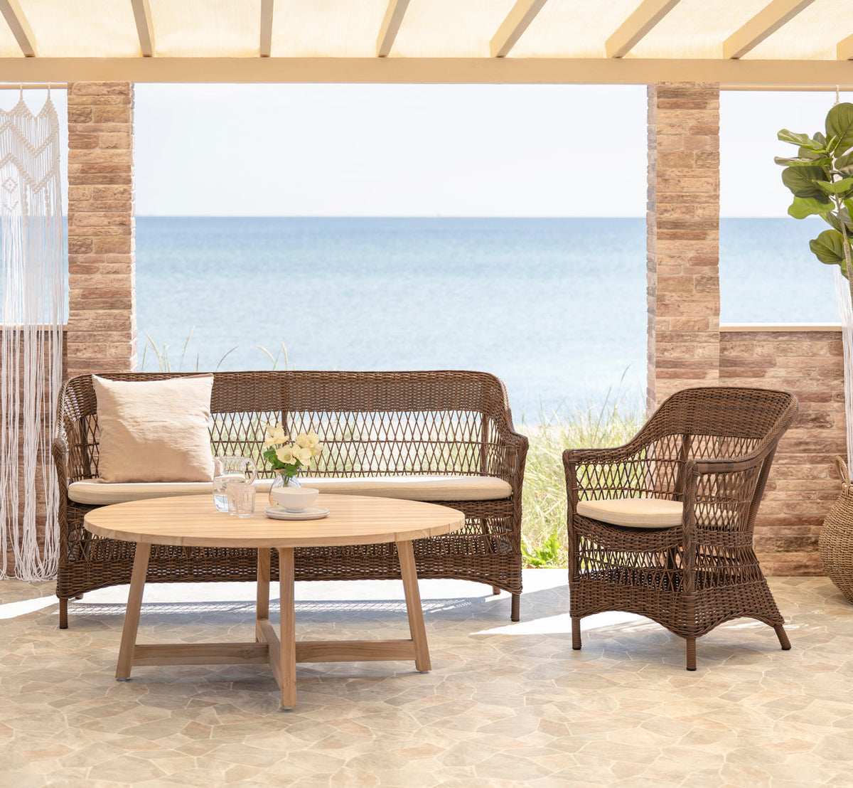 This is a lifestyle image of Charlot Exterior Chair in brown artfibre aluminum. With teak coffee table on a seaside terrace.