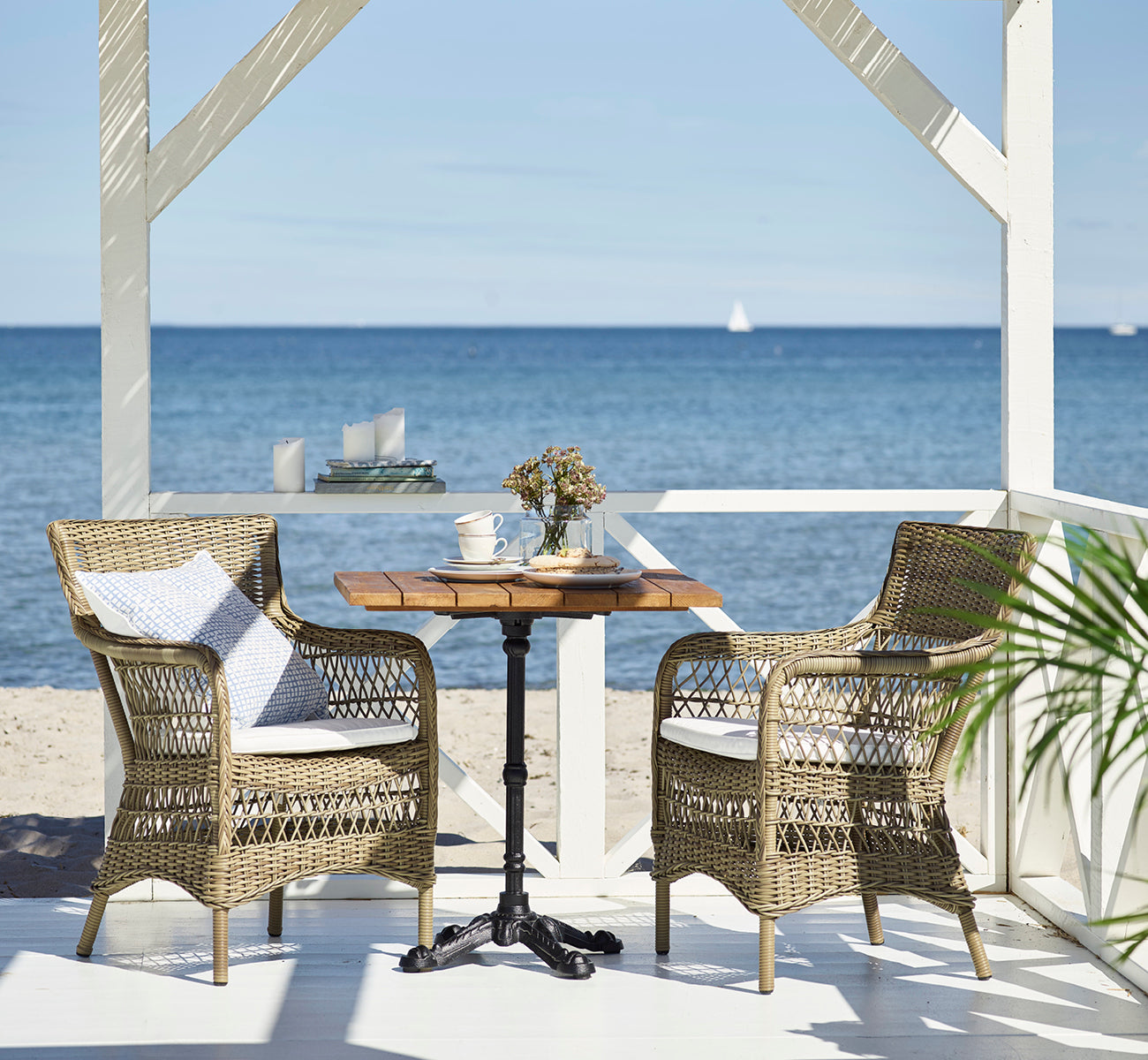 This is a lifestyle image of Marie Exterior Armchair in antique grey artfibre aluminium. At a teak table on a beach pavilion.
