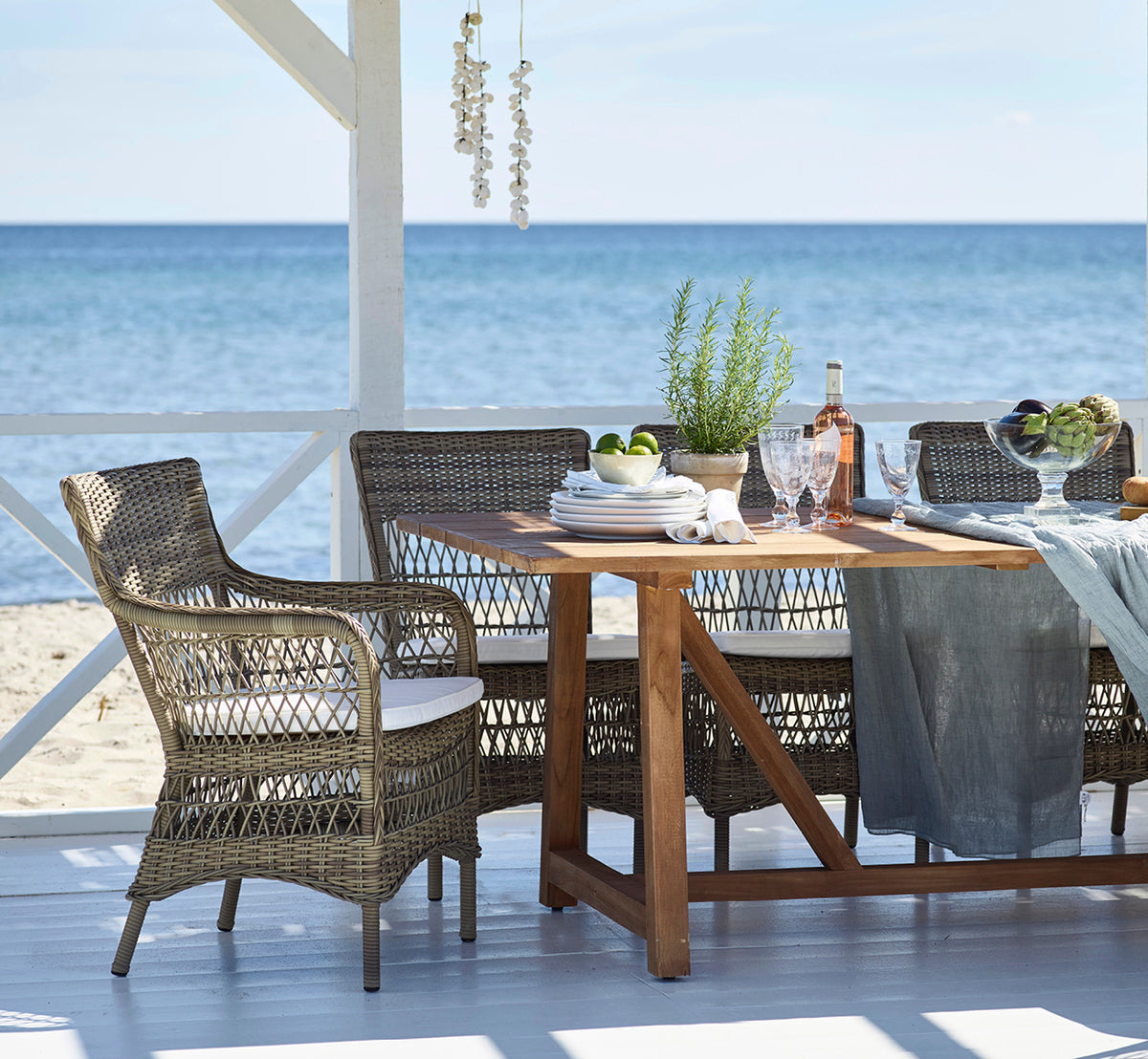 This is a lifestyle image of Marie Exterior Armchair in antique grey artfibre aluminium. Around a teak table at a beach pavilion.