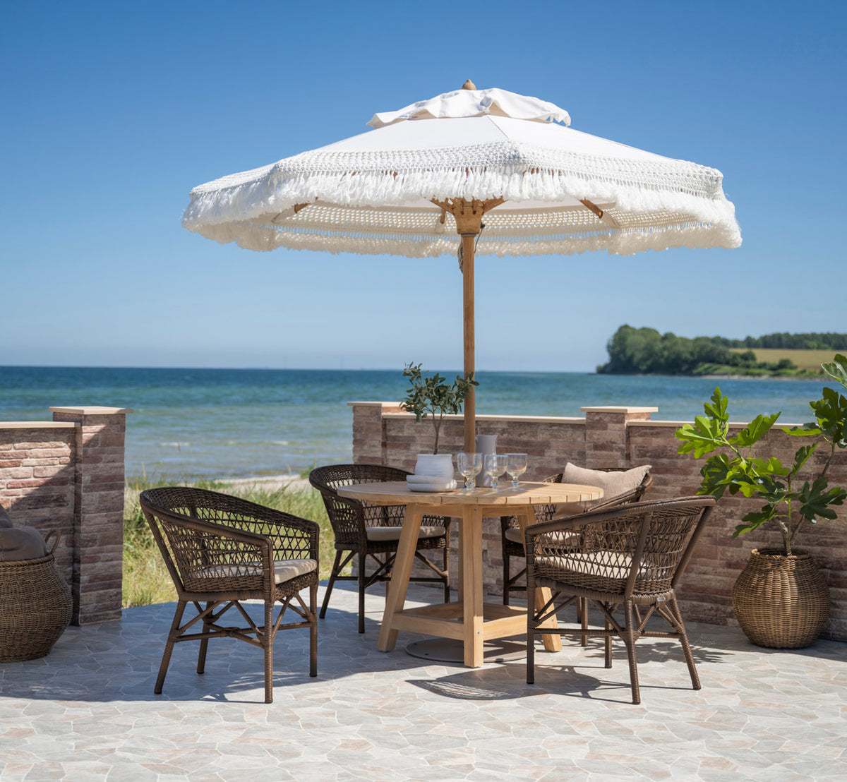 This is a lifestyle image of Emma Exterior Chair in brown artfibre aluminum. Dining under a fringed parasol by the sea.