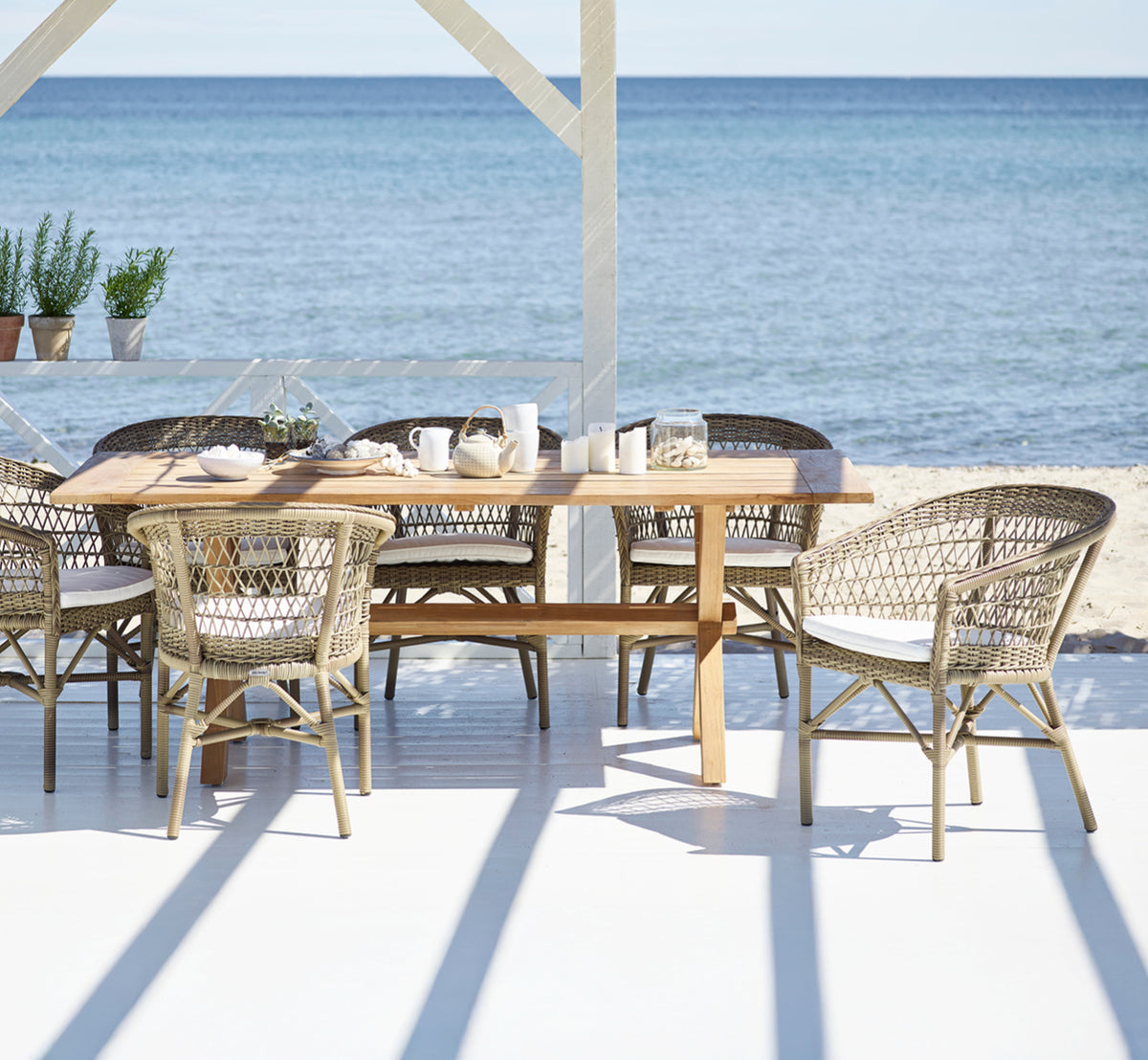 This is a lifestyle image of Emma Exterior Dining Chair in antique Grey ArtFibre Aluminium, on a bright coastal terrace with a reed roof shade and.