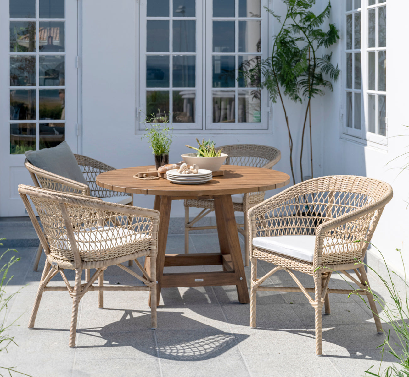 This is a lifestyle image of Emma Exterior Dining Chair in natural ArtFibre Aluminium, on a bright coastal terrace with a reed roof shade and sea.