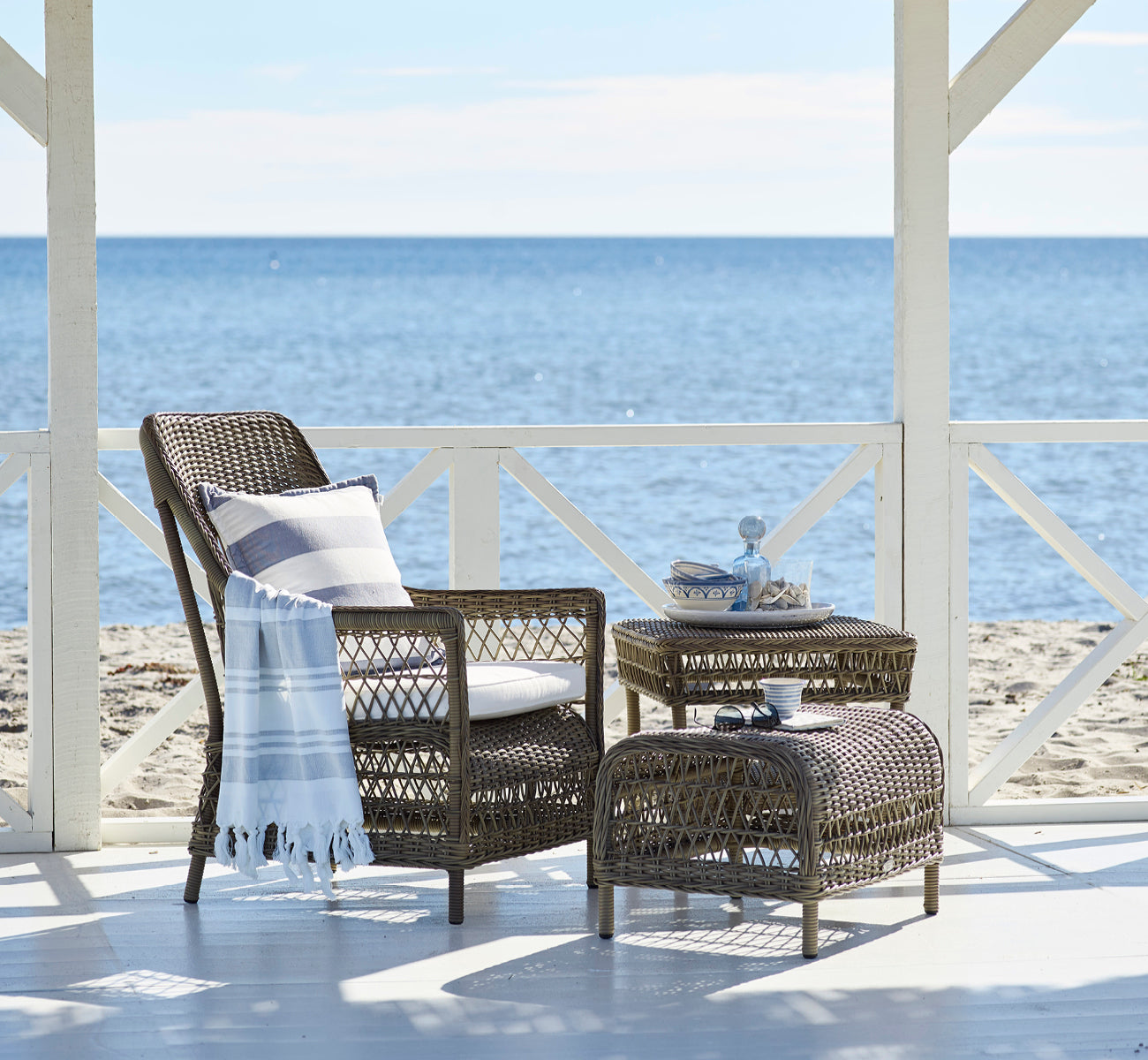 This is a lifestyle image of Dawn Footstool in antique Grey ArtFibre Aluminium, on a coastal terrace with sea views, beach grasses and white columns.