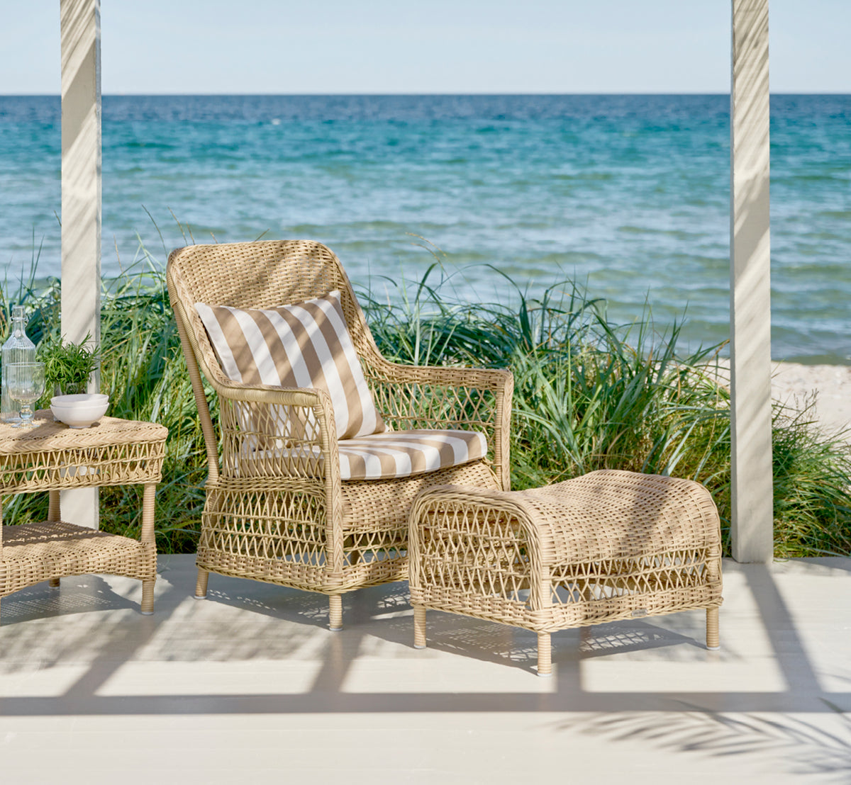 This is a lifestyle image of Dawn Footstool in natural ArtFibre Aluminium, on a coastal terrace with sea views, beach grasses and white columns.