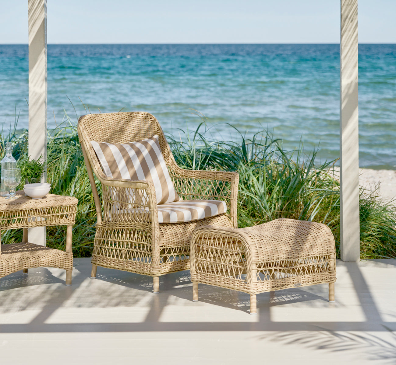 This is a lifestyle image of Dawn Footstool in natural ArtFibre Aluminium, on a coastal terrace with sea views, beach grasses and white columns.