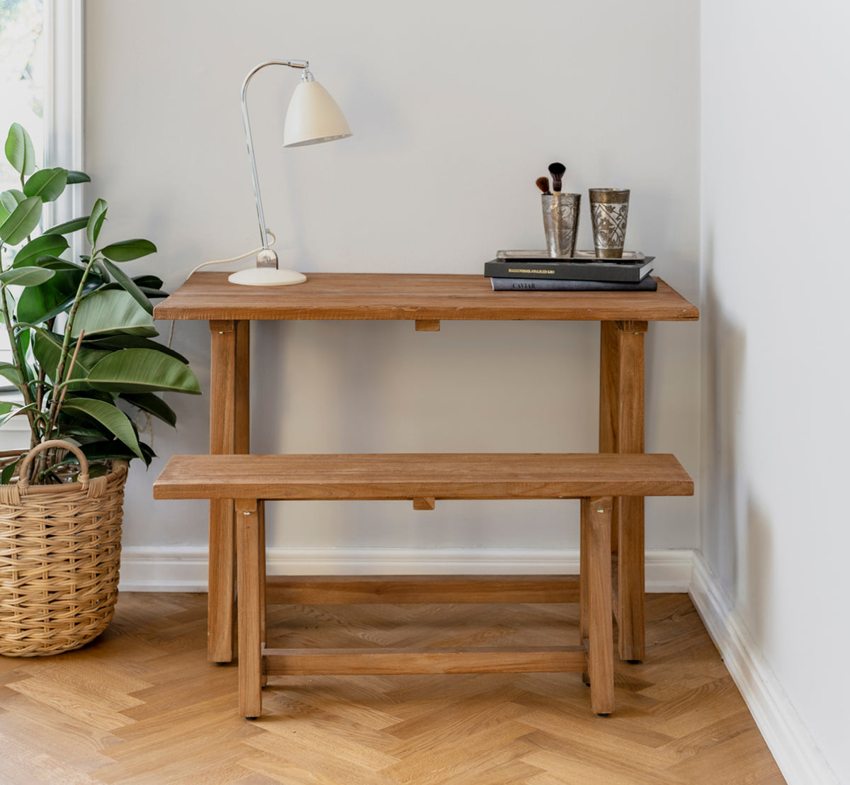 This is a lifestyle image of Lucas Teak Bench in teak indoor. Paired with a teak desk, lamp, and books in a cozy corner on herringbone floor.