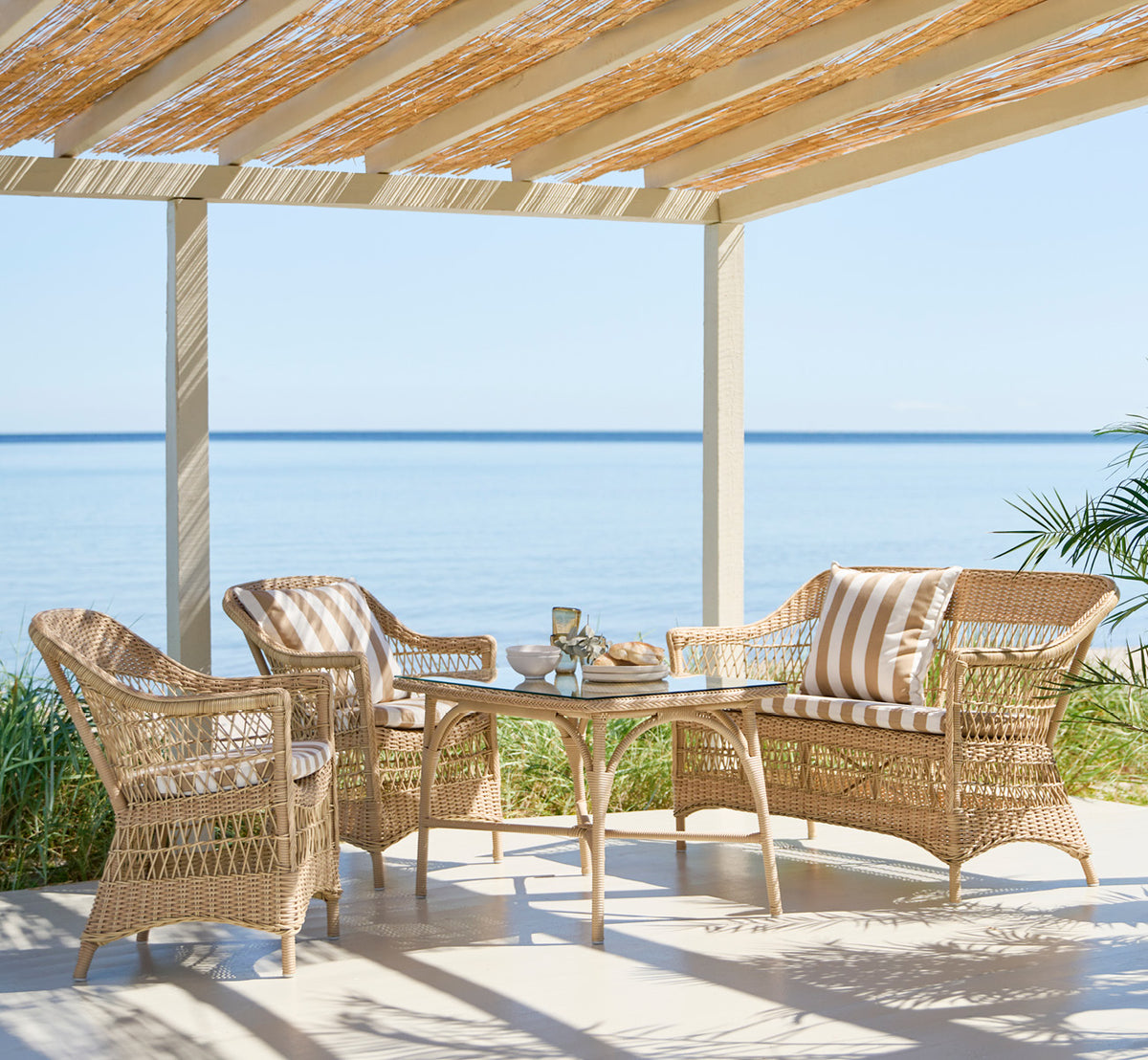 This is a lifestyle image of Charlot 2 Seater Sofa in natural ArtFibre Aluminium, on a coastal terrace with reed roof, sea views and striped cushions.