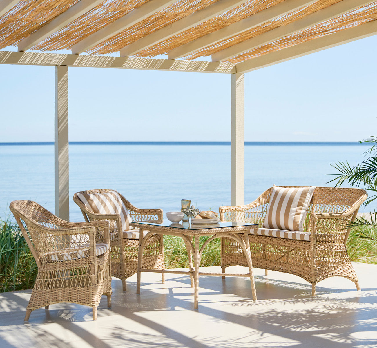 This is a lifestyle image of Charlot 2 Seater Sofa in natural ArtFibre Aluminium, on a coastal terrace with reed roof, sea views and striped cushions.