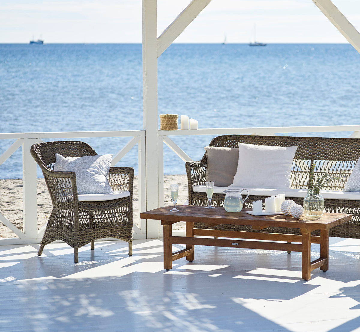 This is a lifestyle image of Charlot 3 Seater Sofa in antique Grey ArtFibre Aluminium, on a coastal terrace with reed roof shade and sea views.