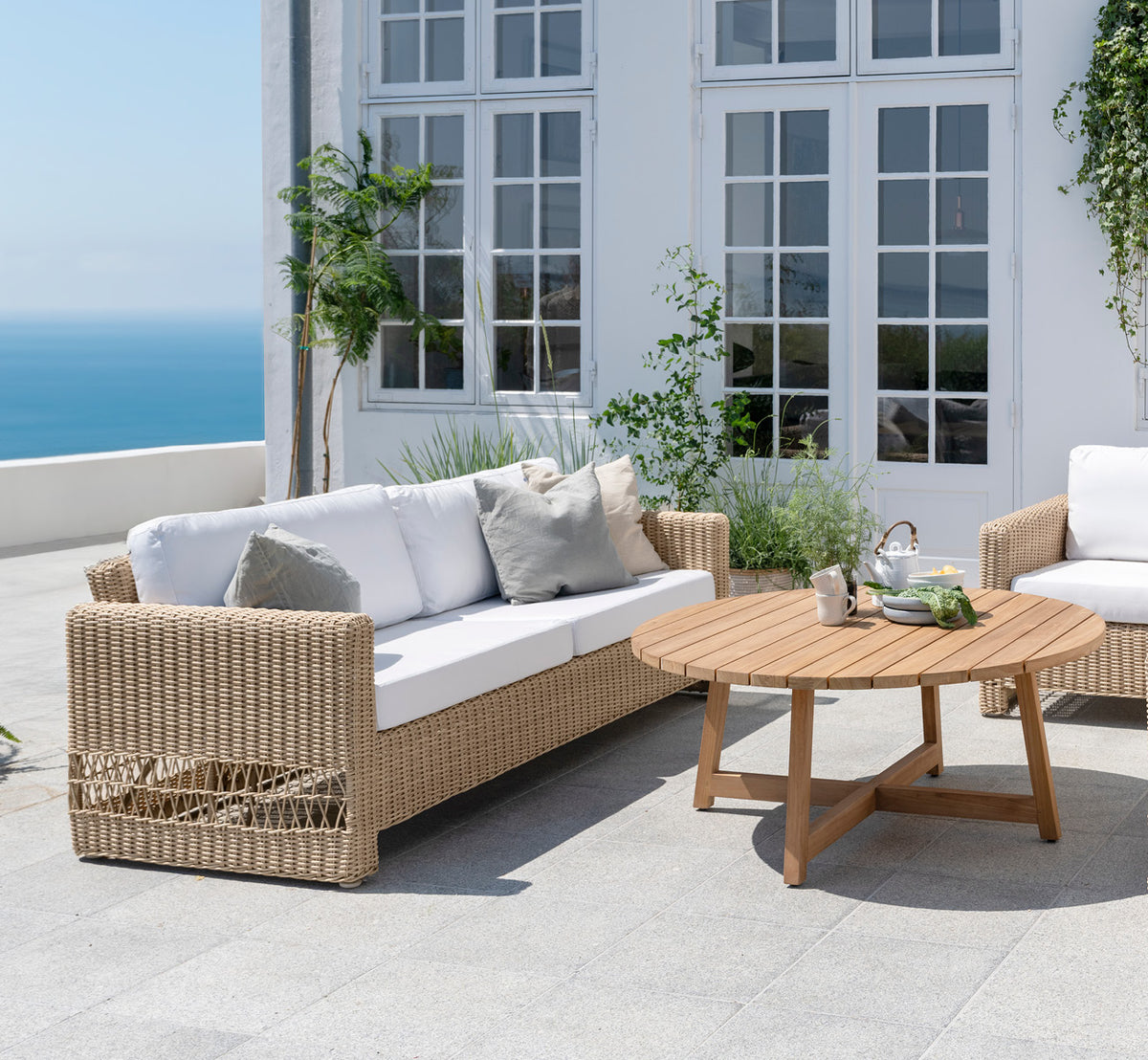 This is a lifestyle image of Carrie Sofa in natural ArtFibre Aluminium, on a coastal terrace with white wooden deck, sea views and pergola.