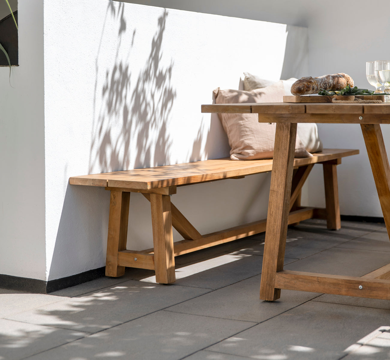 This is a lifestyle image of George Exterior Bench in outdoor Teak, on an outdoor patio with grey tile flooring and a white rendered wall.