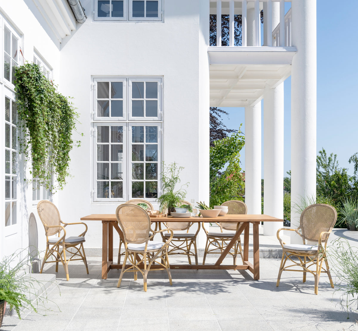 This is a lifestyle image of George Exterior Table 240 in outdoor Teak, on an outdoor garden patio with a white house facade and greenery.