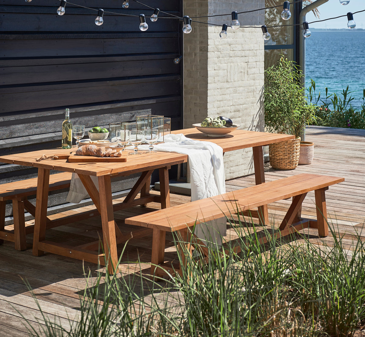 This is a lifestyle image of George Exterior Table 240 in outdoor Teak, on an outdoor garden patio with a white house facade and greenery.