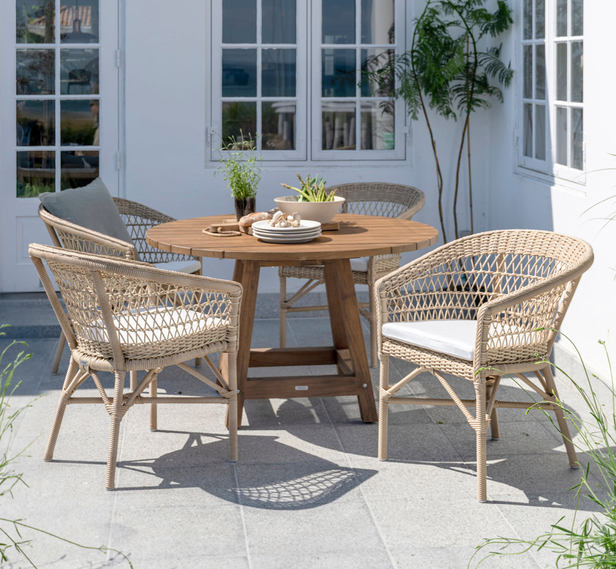 This is a lifestyle image of George Exterior Table Ø120 in outdoor Teak, on an outdoor garden patio with a white house facade and greenery.