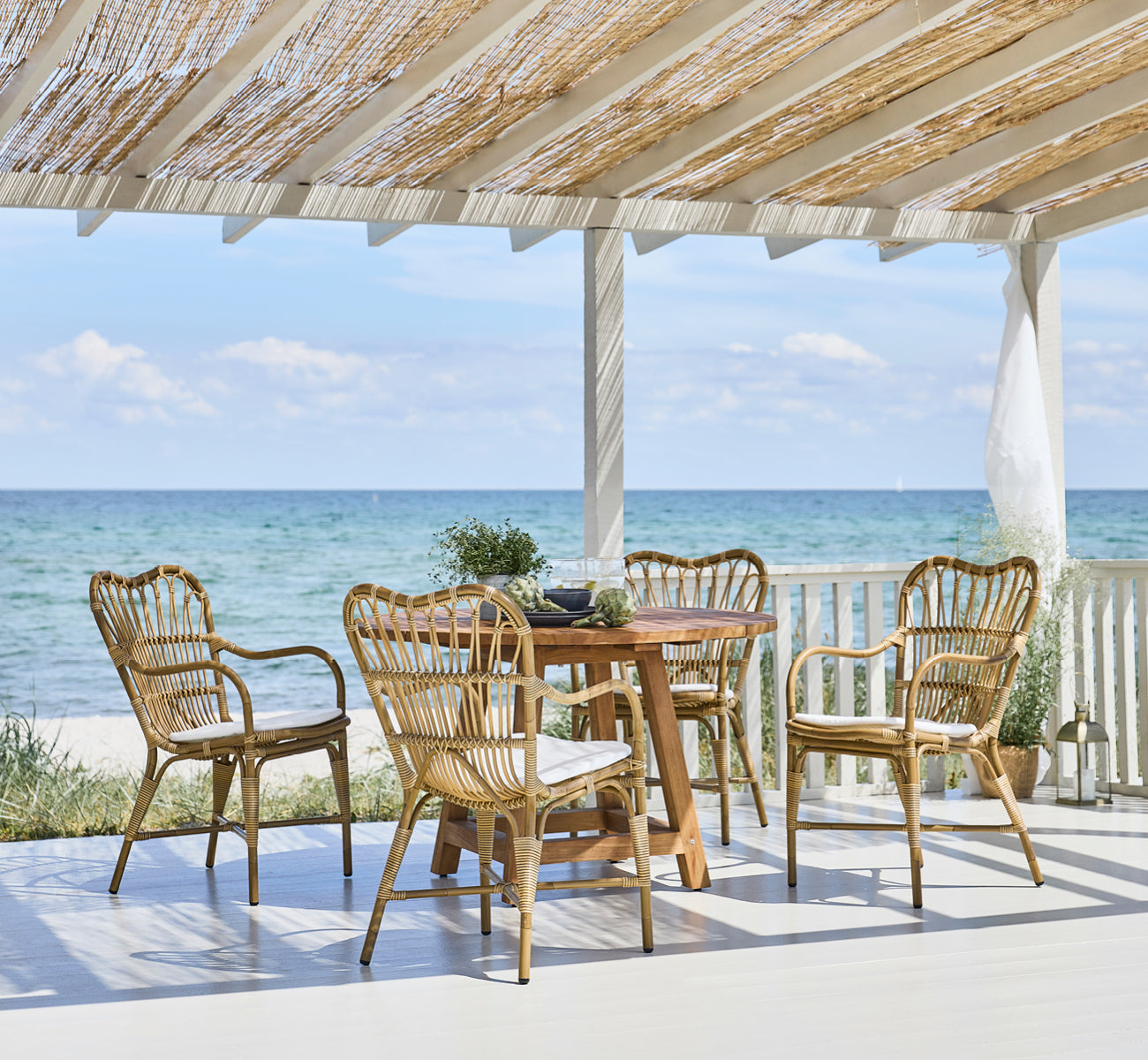 This is a lifestyle image of George Exterior Table Ø120 in outdoor Teak, on an outdoor garden patio with a white house facade and greenery.