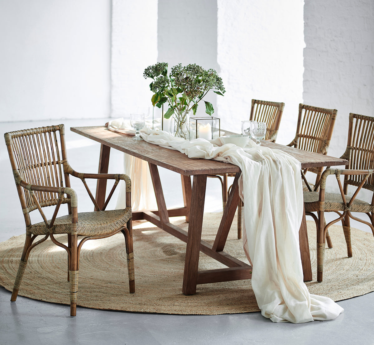This is a lifestyle image of Lucas Teak Dining Table 240X100 cm in teak indoor. Draped with linen and greenery, surrounded by rattan chairs.