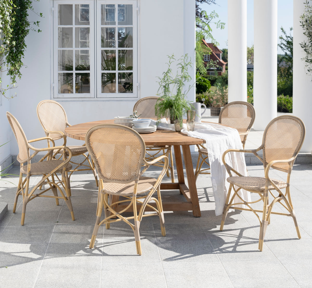 This is a lifestyle image of George Exterior Table Ø160 in outdoor Teak, on an outdoor garden patio with a white house facade and greenery.