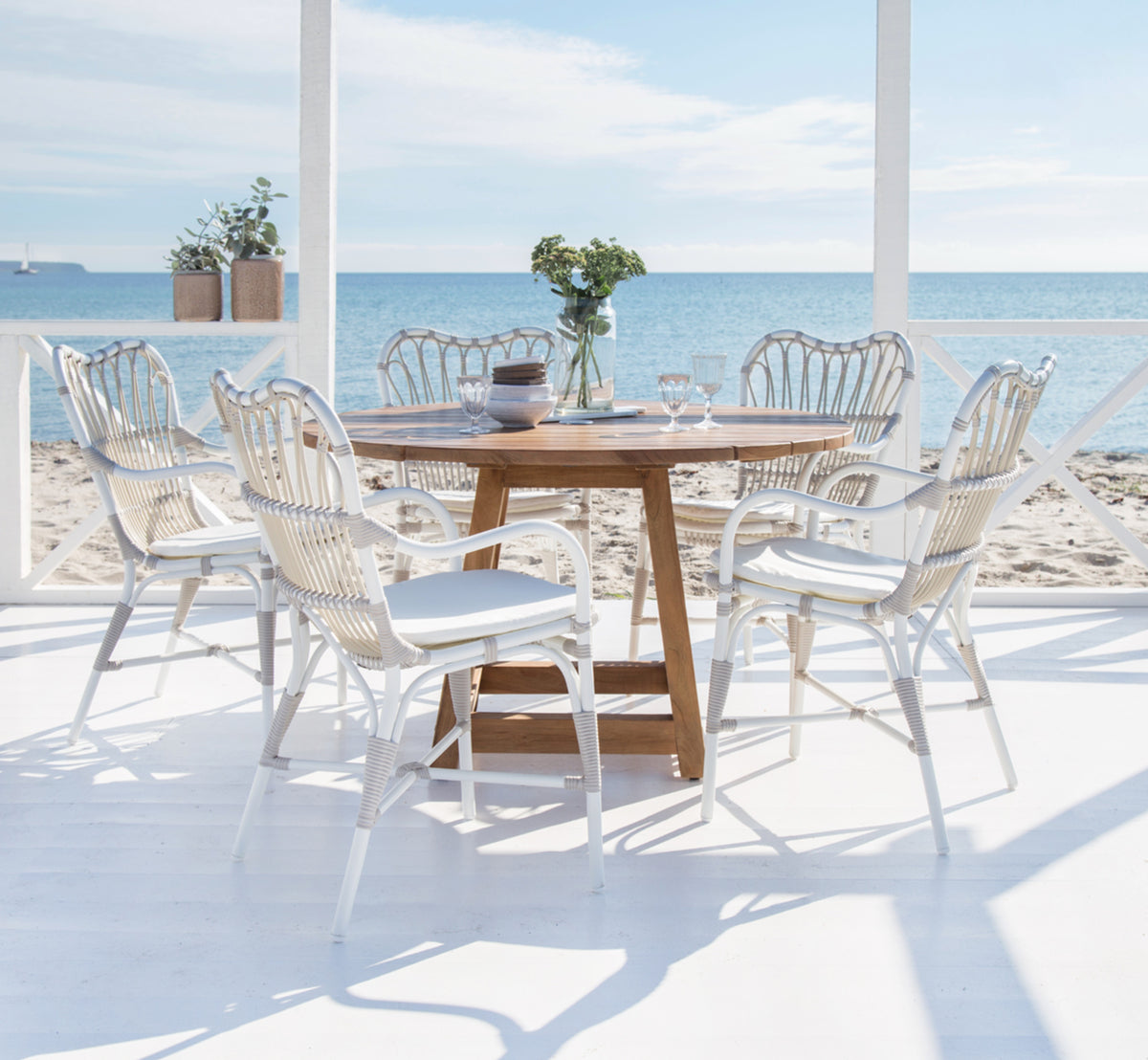 This is a lifestyle image of George Exterior Table Ø160 in outdoor Teak, on an outdoor garden patio with a white house facade and greenery.