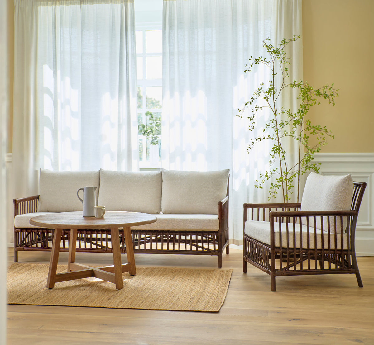This is a lifestyle image of Aksel Coffee Table Ø100 cm in teak indoor. With rattan sofa and lounge chair in a bright room.