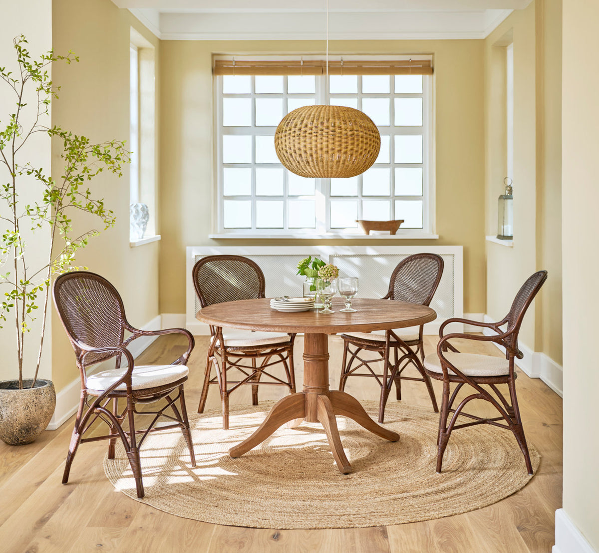 This is a lifestyle image of Michel Teak Dining Table Ø120 cm in teak indoor. With rattan chairs and pendant in a sunny dining room.