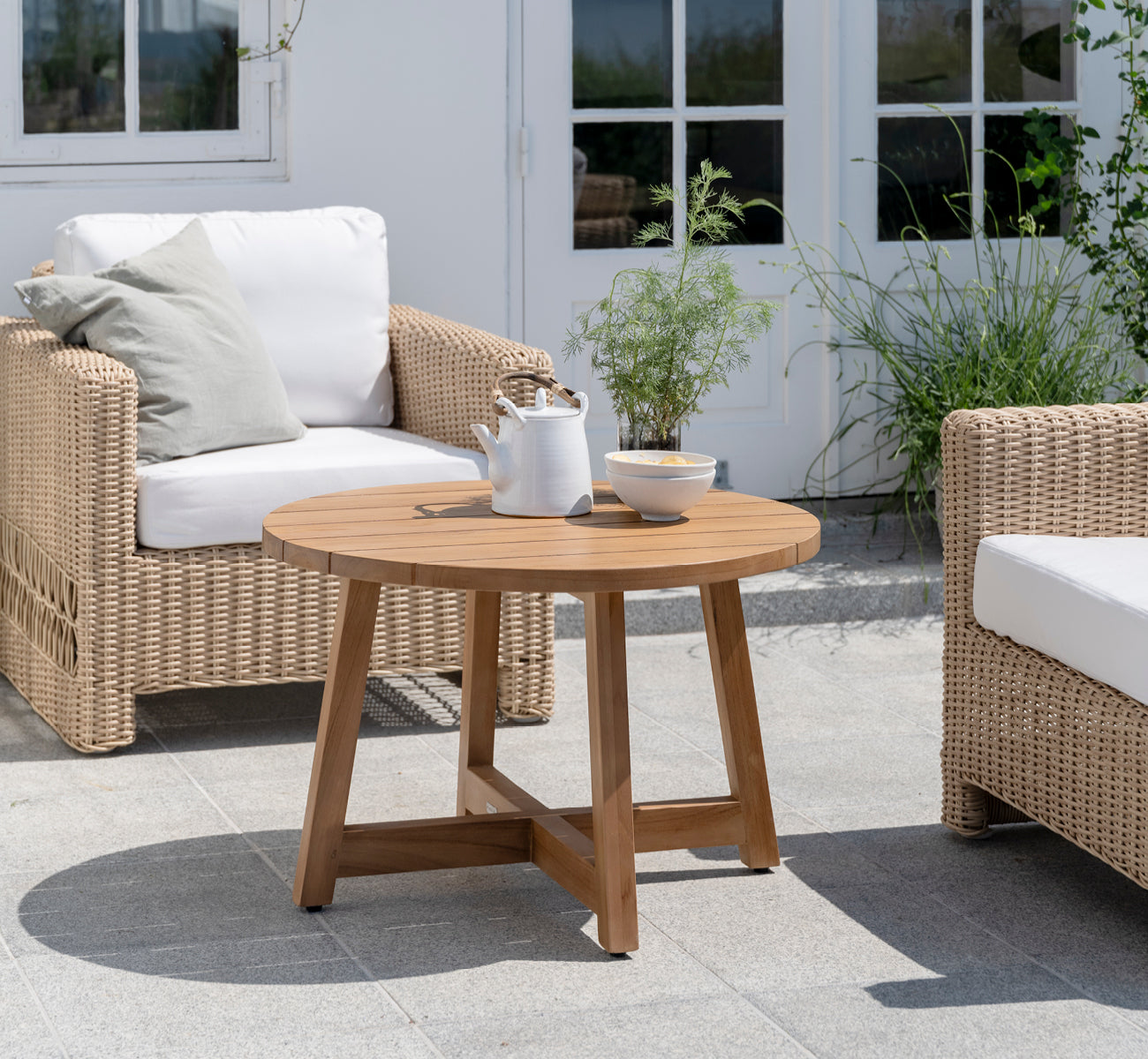 This is a lifestyle image of Victor Teak Lounge Table Ø75 cm in teak outdoor. Set on a seaside terrace between daybeds with striped cushions.