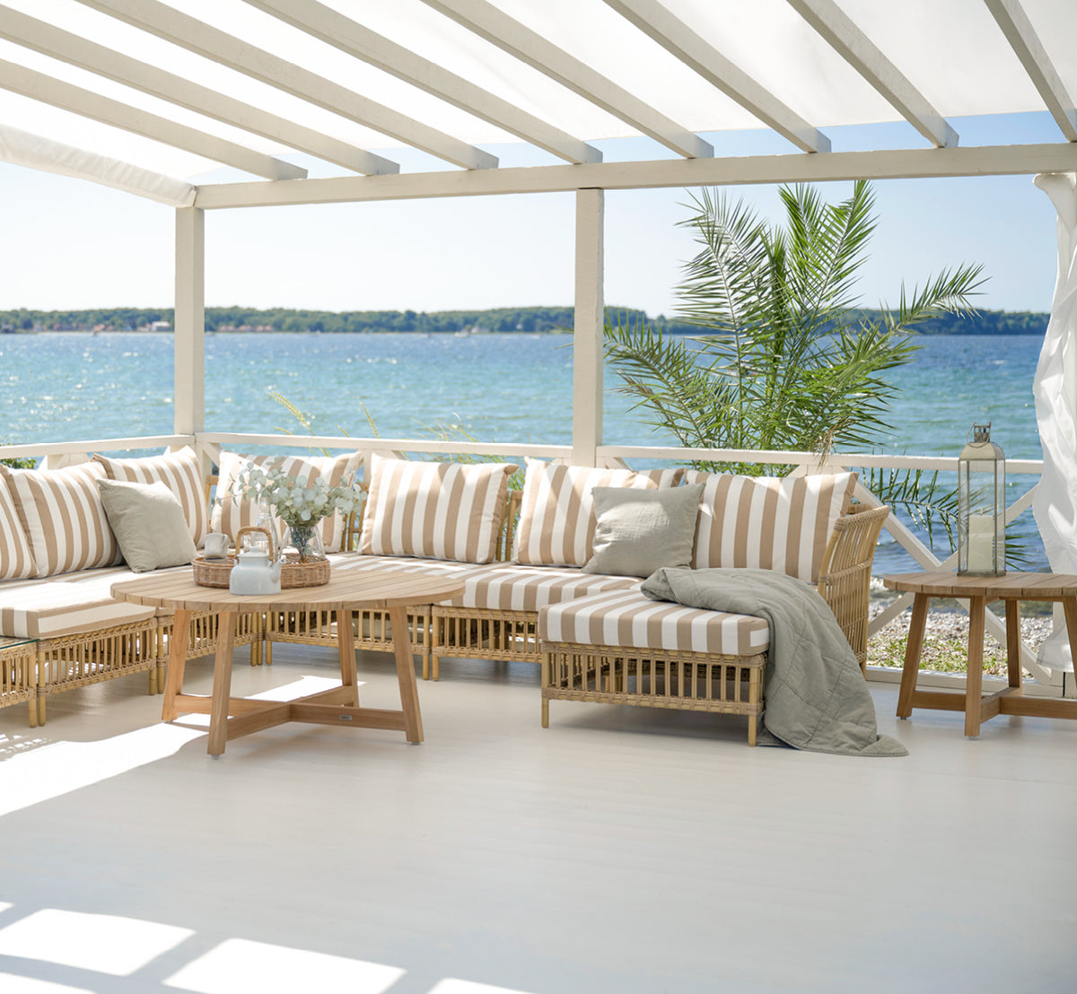 This is a lifestyle image of Victor Teak Lounge Table Ø75 cm in teak outdoor. Set on a seaside terrace between daybeds with striped cushions.
