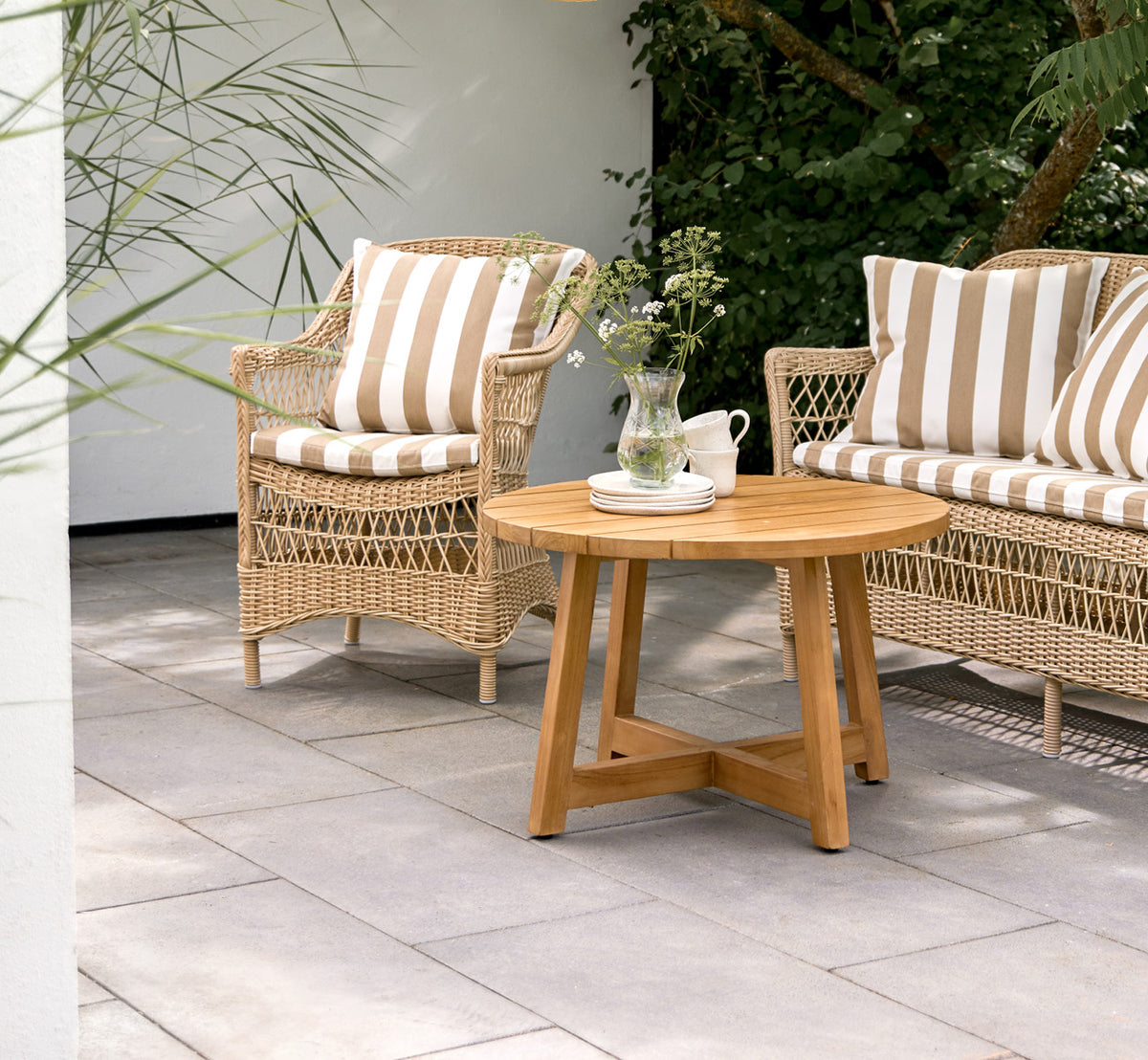 This is a lifestyle image of Victor Teak Lounge Table Ø75 cm in teak outdoor. Set on a seaside terrace between daybeds with striped cushions.