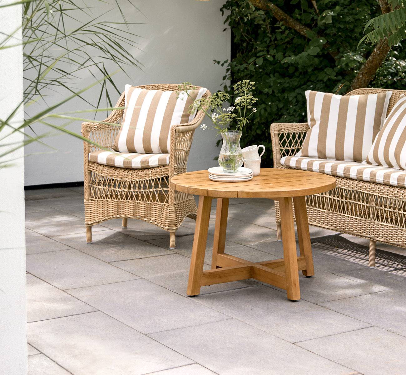 This is a lifestyle image of Victor Teak Lounge Table Ø75 cm in teak outdoor. Set on a seaside terrace between daybeds with striped cushions.