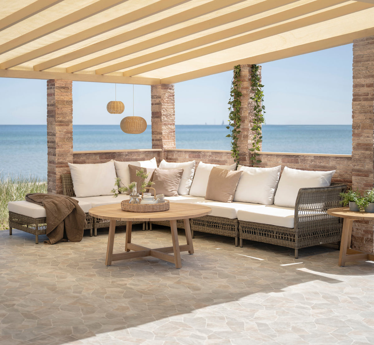 This is a lifestyle image of Victor Teak Lounge Table Ø75 cm in teak outdoor. Corner sofa, pouf and teak table under a seaside pergola.